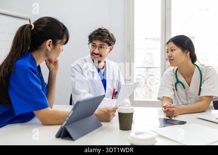 Un team medico si riunisce in una luminosa stanza d'ospedale, collaborando alla cura dei pazienti. Un'infermiera in abito bianco ascolta intensamente mentre due medici discutono del trattamento Foto Stock