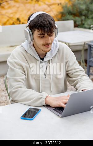 Un giovane uomo siede all'aperto, indossa le cuffie e lavora su un notebook. Accanto a lui c'è uno smartphone sul tavolo. Questa scena cattura l'essenza del mod Foto Stock
