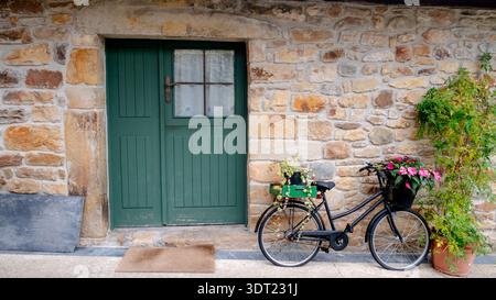 Una bicicletta d'epoca adornata da fiori vibranti si appoggia su un muro rustico in pietra con una porta verde a Tazones, Asturie, Spagna, creando un pittoresco Foto Stock