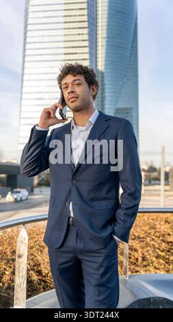 Un uomo d'affari sicuro di sé con una tuta marina si trova all'esterno, impegnato in una conversazione telefonica sullo sfondo di un moderno grattacielo cittadino, che simboleggia la u Foto Stock