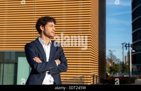 Un giovane professionista in un abito scuro si erge con sicurezza con le braccia incrociate fuori da un edificio moderno, simboleggiando ambizione e successo in un contemporaneo Foto Stock