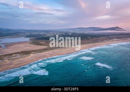 Vista aerea della tranquilla spiaggia costiera e delle onde oceaniche al tramonto pastello sulle dune e il villaggio, Valdovino, Spagna Foto Stock