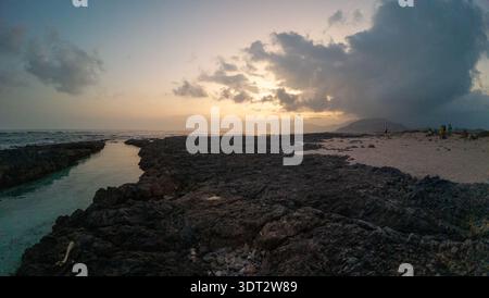 Suggestivo tramonto costiero sulla costa rocciosa con canale di marea, cielo nuvoloso, collina lontana e piccole sagome sulla spiaggia sabbiosa; tranquillo paesaggio marino Foto Stock