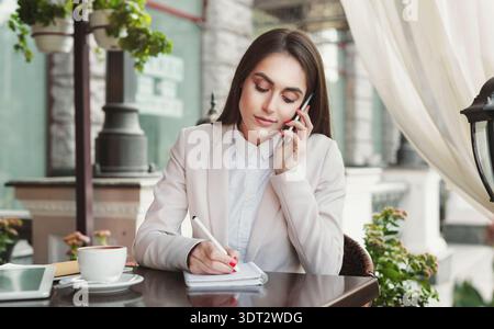 Donna d'affari che prende appunti durante una telefonata in un caffè durante il giorno Foto Stock