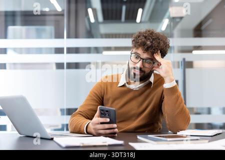 Un giovane con i capelli ricci e la barba si siede a una scrivania in ufficio tenendo in mano uno smartphone, sembra annoiato e confuso, illustrando procrastinazione e distrazione Foto Stock