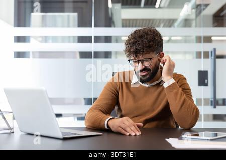 Uomo con capelli ricci e occhiali che soffre di forti dolori alle orecchie e mal di testa, toccando l'orecchio ed esprimendo disagio mentre lavora con un notebook in una Foto Stock