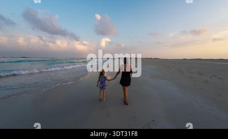 Madre e figlia che si tengono per mano camminando lungo una tranquilla spiaggia di sabbia al tramonto, onde dolci e cielo pastello, ampio paesaggio costiero con copia spa Foto Stock