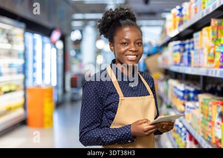 Il lavoratore della drogheria in grembiule sorride alla fotocamera mentre utilizza un tablet digitale per gestire l'inventario e assistere i clienti in un ambiente di corridoio moderno del supermercato Foto Stock