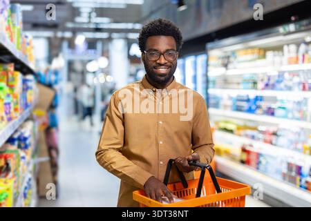 Uomo afroamericano sorridente alla macchina fotografica mentre tiene in mano un cestino arancione e ci mette gli oggetti, in piedi in un corridoio luminoso del supermercato con scaffali di prodotti Foto Stock