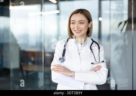 Giovane donna sicura di sé medico in piedi con le braccia incrociate, con indosso un camice da laboratorio bianco e uno stetoscopio, sorridente e guardando lontano in una moderna clinica medica o in un ambiente di ufficio Foto Stock