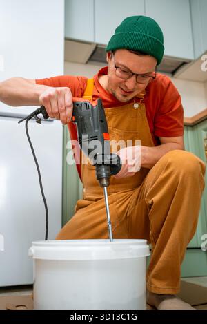 Artigiano maschio che mescola un adesivo bianco per Malta o piastrelle con un miscelatore per trapano in un secchio di plastica, preparando materiali per miglioramenti domestici e lavori di riparazione professionali Foto Stock