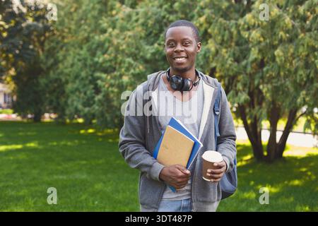 Young Man che tiene in mano caffè e libri nel parco durante il pomeriggio di sole Foto Stock