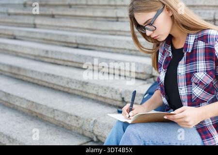 Una giovane donna scrive su notebook mentre è seduta su passi concreti all'aperto, alla luce del giorno Foto Stock