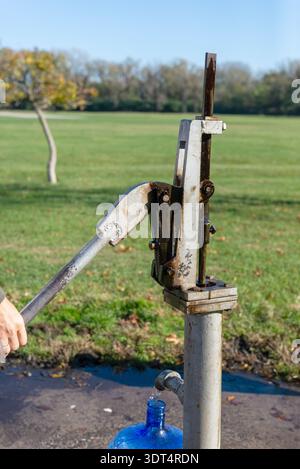 Un primo piano verticale di una pompa dell'acqua manuale del ferro in funzione per riempire una grande caraffa blu. Lo sfondo presenta un campo verde sfocato e alberi Foto Stock