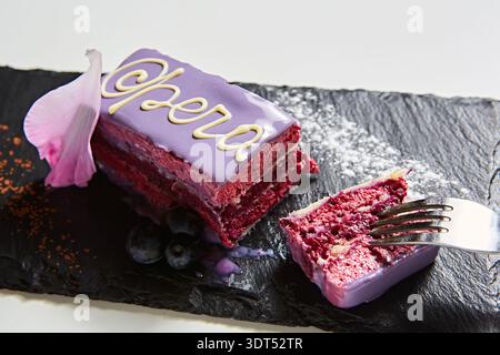 Moderna fetta di torta Opera con vibrante glassa viola, delicata calligrafia al cioccolato bianco e mirtilli freschi, un dessert multi-sensoriale che mette in risalto Foto Stock