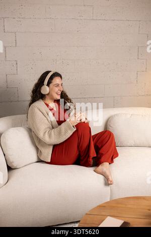 Giovane donna che sorride, indossa le cuffie e siede comodamente sul divano di casa Foto Stock