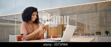 Giovane donna d'affari africana felice che usa il telefono cellulare seduto in ufficio. Foto Stock