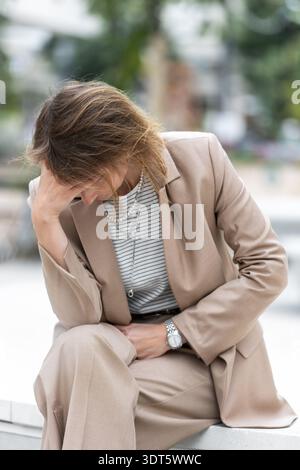 Disperazione donna d'affari esausta indossa un abito beige seduto all'aperto panca urbana testa in mano stress ansia burnout crisi mentale. Pensivo di mezza età Foto Stock