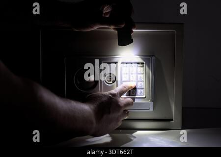 Uomo che inserisce il codice di sicurezza sulla cassetta di sicurezza elettronica in una stanza buia con torcia. Concetto di mistero, risparmi nascosti, sicurezza domestica e protezione finanziaria. Foto Stock