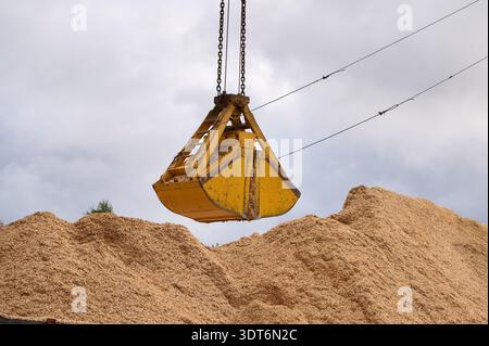 I macchinari pesanti sollevano la benna su un mucchio di trucioli di legno in un cantiere nel pomeriggio Foto Stock