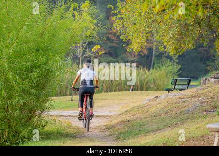 Un ciclista pedala su una bicicletta rossa lungo un sentiero sterrato attraverso il verde lussureggiante e il giallo fogliame autunnale in un tranquillo parco. Foto Stock