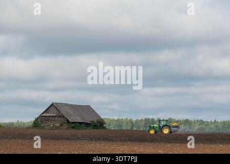 Trattore verde che spande fertilizzante su un campo appena arato accanto a un vecchio fienile di legno. La scena agricola rurale che simboleggia l'agroalimentare e la produzione di colture, Foto Stock