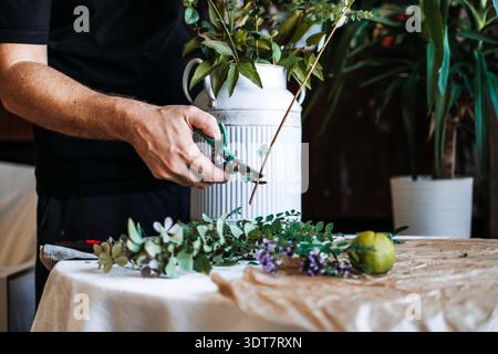 Le mani utilizzano cesoie verdi da giardino per rifinire il sottile stelo botanico sul tavolo accanto alla caraffa scanalata bianca. biofilia, vita ispirata alla natura, connessione vegetale-umana Foto Stock