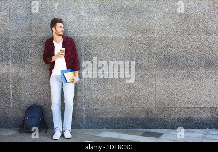 Young Man si trova accanto al muro mentre tiene in mano caffè e libri nell'area urbana Foto Stock