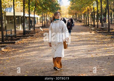 Donna che cammina attraverso il vicolo autunnale del parco con indosso un lungo cappotto bianco Foto Stock