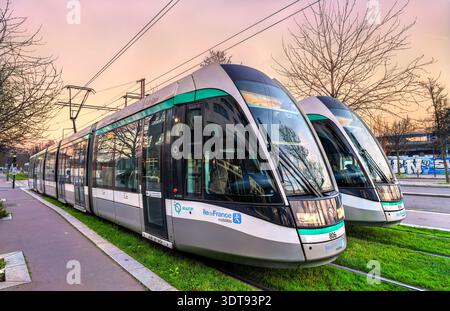 Saint Denis, Francia - 29 dicembre 2025: Due moderni tram passeggeri Alstom su piste di erba verde al tramonto. I mezzi di trasporto pubblico operano vicino a Parigi. Foto Stock
