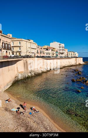 Le persone trascorrono del tempo sulla spiaggia di Siracusa Ortigia, godendosi il sole e l'acqua. Il litorale ha acqua limpida e ciottoli. Foto Stock