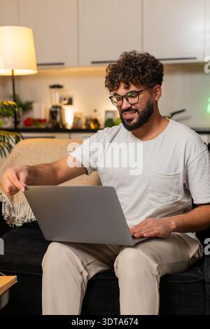 L'uomo arabo a casa apre il laptop lancia i tipi di progetto velocemente funziona sorrisi a distanza concentrati. Il ragazzo egiziano su freelance divano risponde ai file di modifica delle e-mail ch Foto Stock