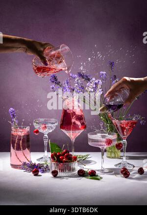 Le mani dei barman versano bevande rosse e viola in bicchieri da cocktail assortiti e creano schizzi, circondati da ciliegie fresche e lavanda Foto Stock