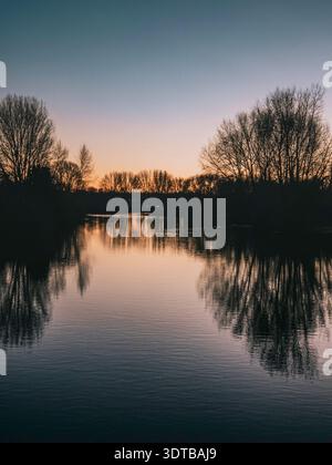 Un tranquillo ambiente invernale caratterizzato da un sentiero ghiacciato che si snoda verso un ponte, illuminato da un'unica lampada. Il cielo passa dal crepuscolo alla notte, W Foto Stock
