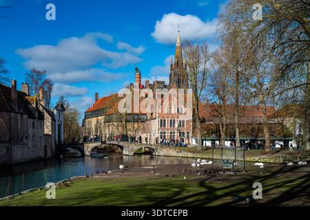 Bruges, Belgio - 14 febbraio 2026: I visitatori attraversano il ponte vicino a Begijnhof e Wijngaardplein con la torre della Chiesa di nostra Signora che sorge in istoria Foto Stock