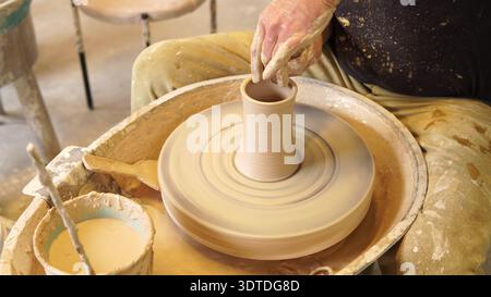 Primo piano sulla sagomatura e la raffinazione di un vaso di argilla alto sulla ruota rotante in un laboratorio di ceramica Foto Stock
