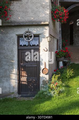 Affascinante entrata rustica da cottage con porta di legno scuro adornata con tende in pizzo e battitori a forma di cuore. Gerani rossi vibranti cadono dalla piantatrice Foto Stock
