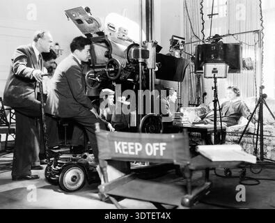 Questa fotografia d'epoca degli anni '1930 cattura un momento dietro le quinte sul set di "l'uomo sottile" nel 1934, con il direttore della fotografia James Wong Howe che gestisce la fotocamera, con l'attrice Myrna Loy seduta su un divano. La scena è illuminata da grandi luci cinematografiche, che mostrano le prime tecniche del cinema hollywoodiano. Foto Stock