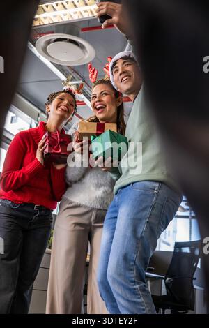 Diversi colleghi che si pongono per selfie in un ufficio aperto con smartphone, regali, corna e cappello di Babbo Natale Foto Stock