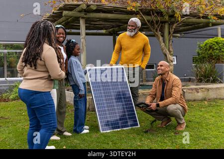 Famiglia afroamericana che prova il pannello solare sul prato, senior steadying, inginocchiato che tiene la tavoletta Foto Stock