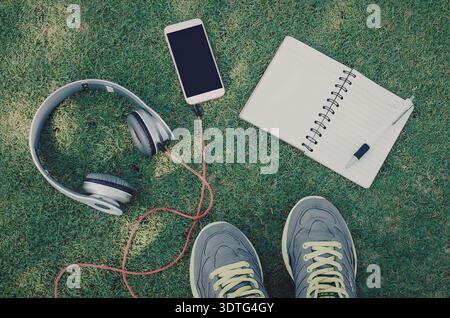I piedi di una persona indossano scarpe da ginnastica accanto a un notebook, una penna, uno smartphone con cuffie su un campo erboso. Foto Stock