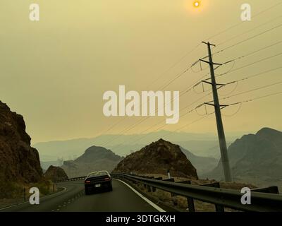 Guida in auto lungo una tortuosa strada desertica vicino alla diga di Hoover con linee elettriche e paesaggi di montagna nebbiosi sotto il cielo giallo in Nevada, Stati Uniti. Foto Stock