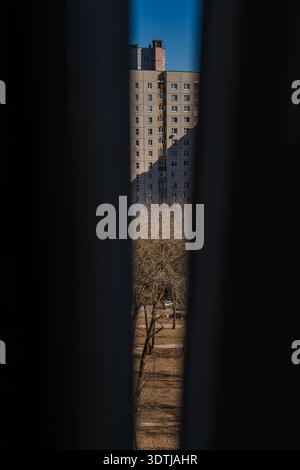 Un alto blocco di pannelli grigio appare attraverso uno stretto spazio, con alberi senza foglie, un piccolo sentiero e un'auto bianca parcheggiata sotto clea nel tardo pomeriggio Foto Stock