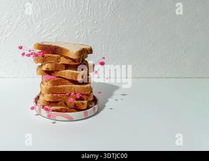 Composizione STILL Life minimalista con fette di pane impilate decorate con piccoli fiori rosa su uno sfondo bianco pulito. Ombreggiatura e copia forti Foto Stock