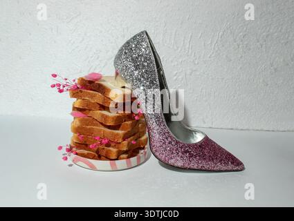 Composizione creativa di Still Life con una scarpa da sospiro glitter rosa posizionata accanto a fette di pane impilate decorate con piccoli fiori rosa. Minimalista con Foto Stock