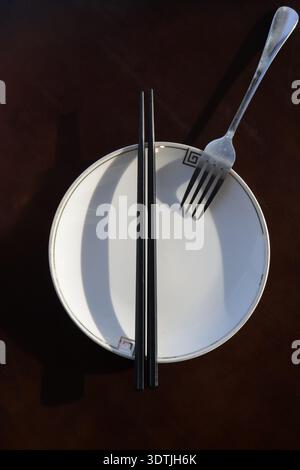 La forca si trova sul lato destro della piastra e le bacchette si trovano sul lato sinistro. Piatto con utensili da cucina, vista dall'alto, su un tavolo rosso scuro. Foto Stock