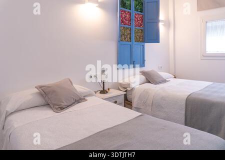 Camera da letto moderna e pulita con due letti singoli con biancheria bianca e getti grigi, con persiane colorate e blu aperte Foto Stock