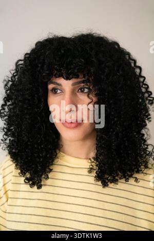 Ritratto di una donna con bellissimi capelli neri ricci che indossa una t-shirt a righe giallo chiaro. Lo sfondo è neutrale, evidenziando il suo occhio espressivo Foto Stock