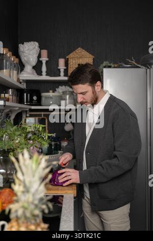 Un uomo con un casual maglione grigio fetta di cavolo rosso su un tagliere di legno in un'accogliente cucina. Gli scaffali pieni di utensili da cucina e vegetazione ne esaltano la qualità Foto Stock