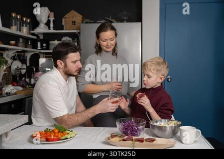 Una famiglia di tre persone si riunisce in cucina per preparare un pasto insieme. Il padre e il figlio si scambiano gli occhiali mentre la madre sorride, circondati da freschi Foto Stock